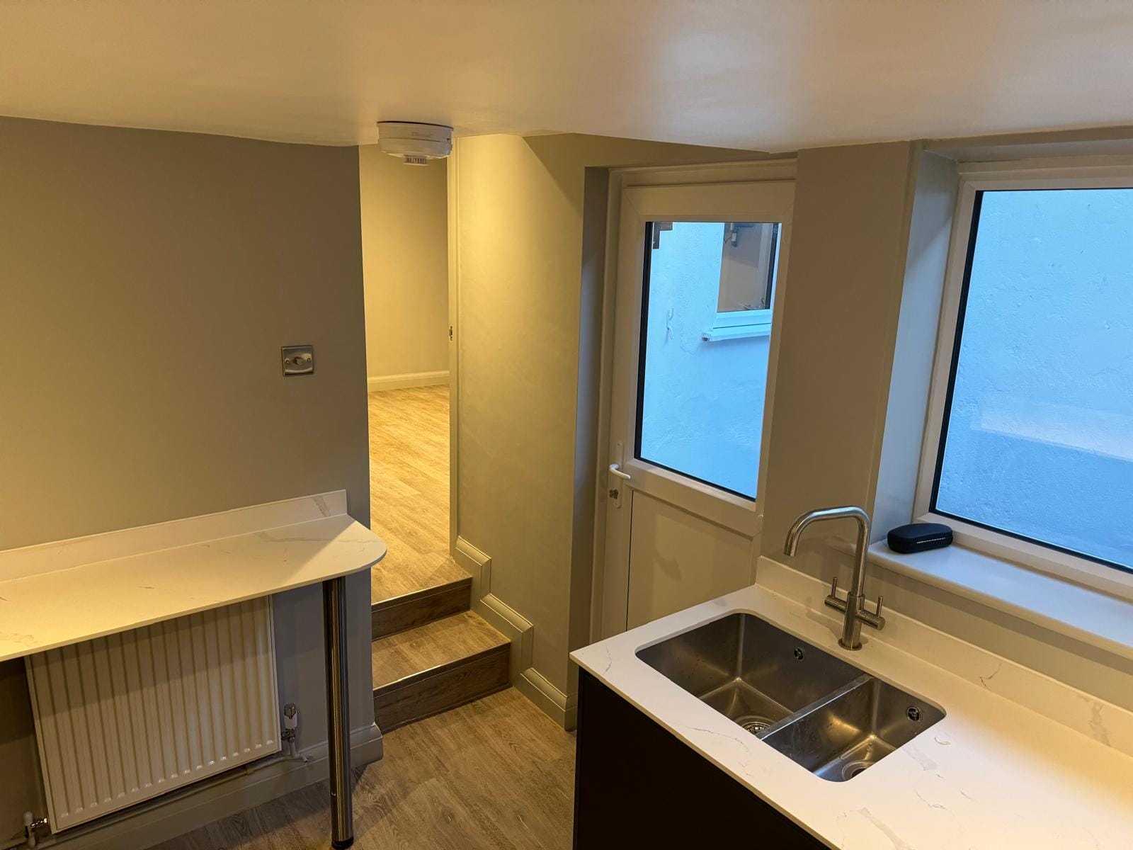 Kitchen with white countertop and double sink, stainless faucet, radiator, and window showing blue exterior wall.