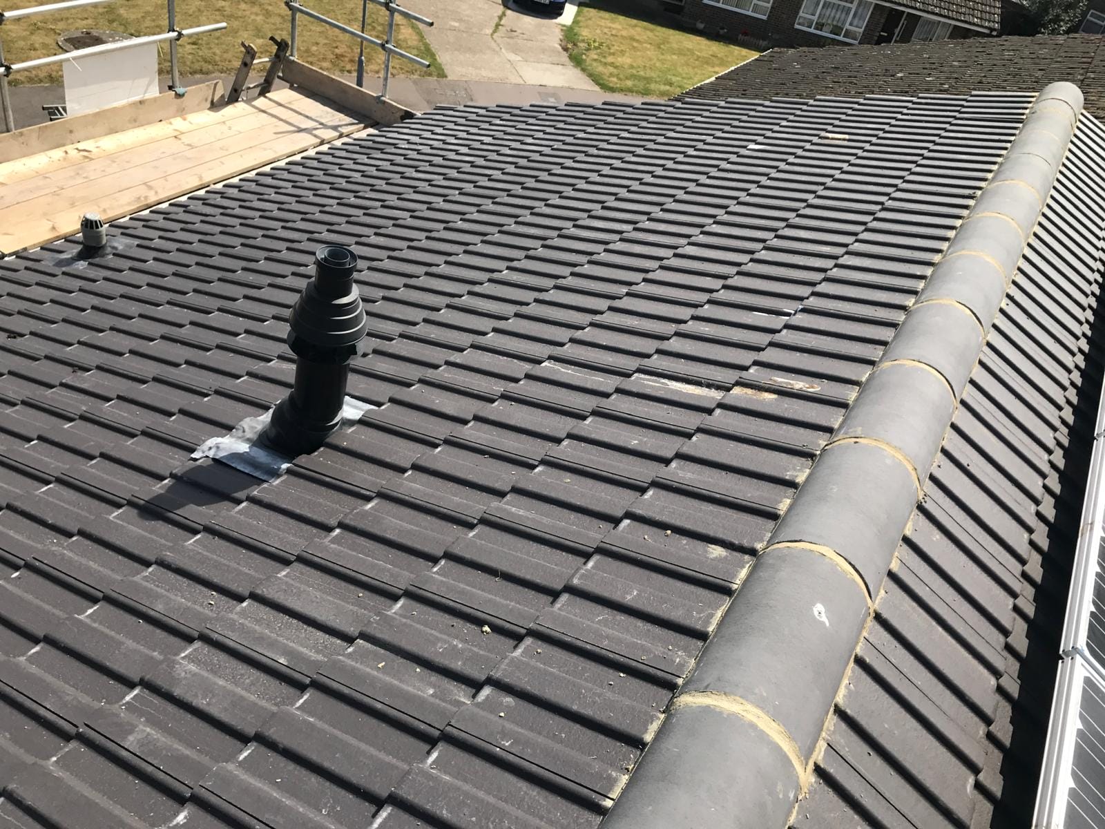 Black tiled roof with a tall vent pipe near the center; temporary scaffolding and a wooden work platform visible at the left edge.